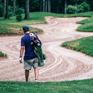 Golfer seen from behind carrying a golf bag over his shoulder beside a winding path lined with grass and trees at Golf de Saint-Roman.