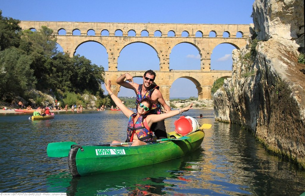 Two people paddling a green canoe on the Gardon River beside rocky cliffs, with the arches of the Pont du Gard in the background.
