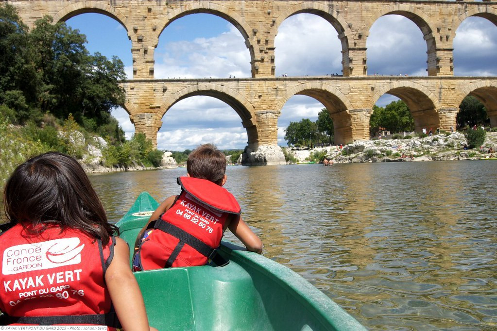 Two children wearing red life jackets in a green canoe on the Gardon River, facing the large arches of the Pont du Gard.
