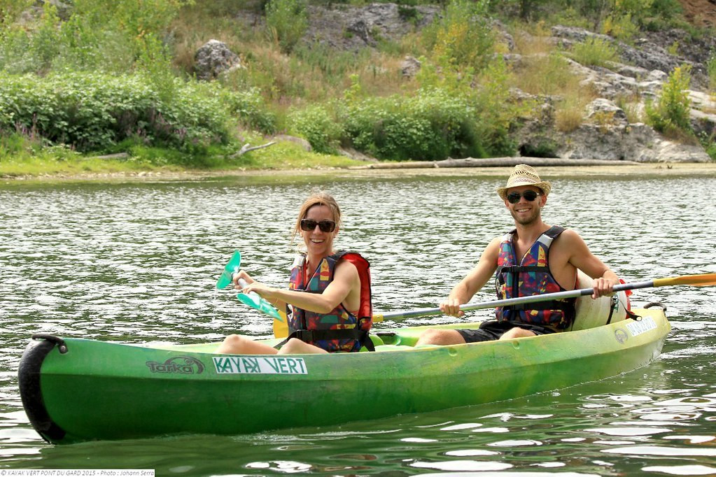 A couple wearing life jackets paddling a green canoe on the calm waters of the Gardon River, with a natural rocky riverbank behind them.