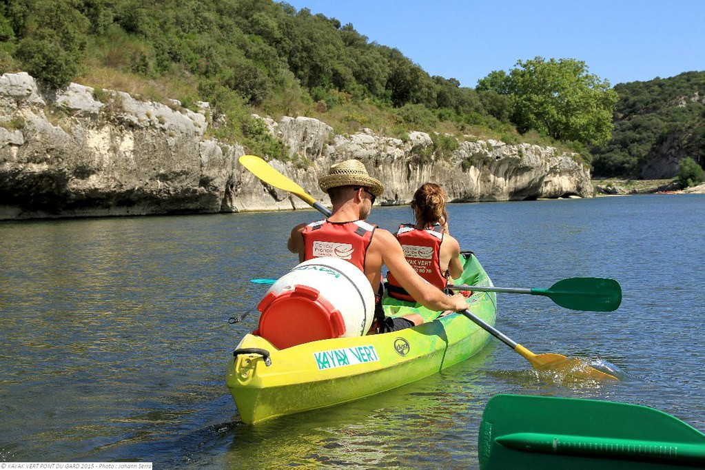 Two people seen from behind paddling a green canoe along the Gardon River, between rocky banks and the greenery of the gorge.