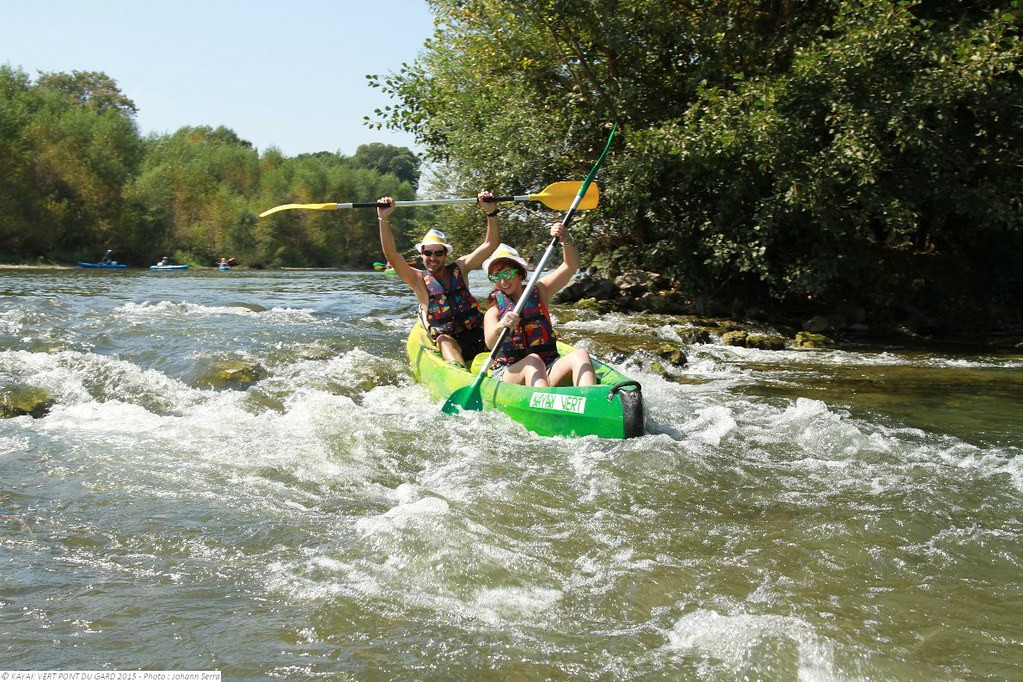 Two people wearing life jackets navigating a stretch of white water in a green canoe on the Gardon River, between tree-lined banks.