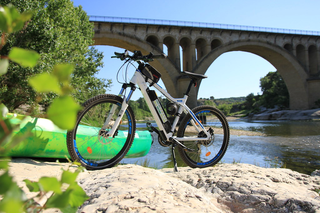 Electric mountain bike resting on a rocky bank of the Gardon River, with a large-arched stone bridge in the background.