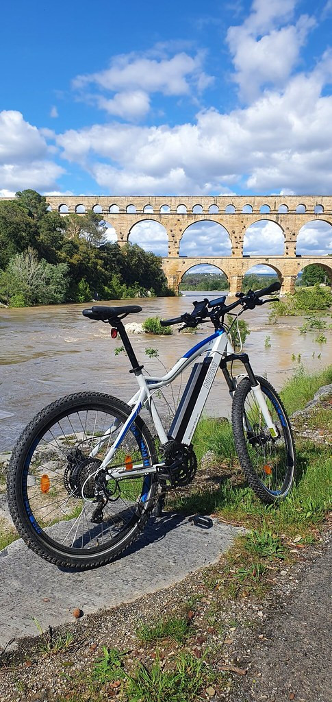 Electric mountain bike parked on the bank of the Gardon River, with the arches of the Pont du Gard in the background beneath a partly cloudy blue sky.