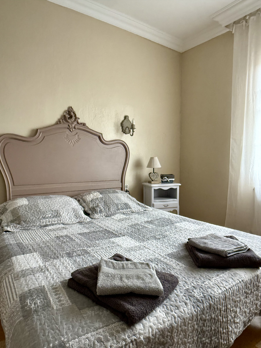 Chambre du gîte Le Nid d’Amour à Vallabrègues avec lit double, tête de lit moulurée, couvre-lit gris, serviettes pliées et fenêtre à rideaux blancs.