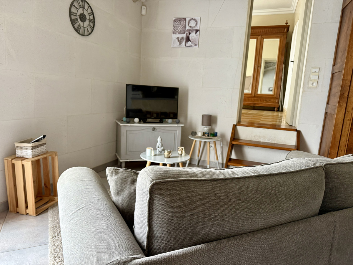 Salon du gîte Le Nid d’Amour avec canapé gris, télévision sur meuble blanc, petite table basse et ouverture vers une autre pièce.