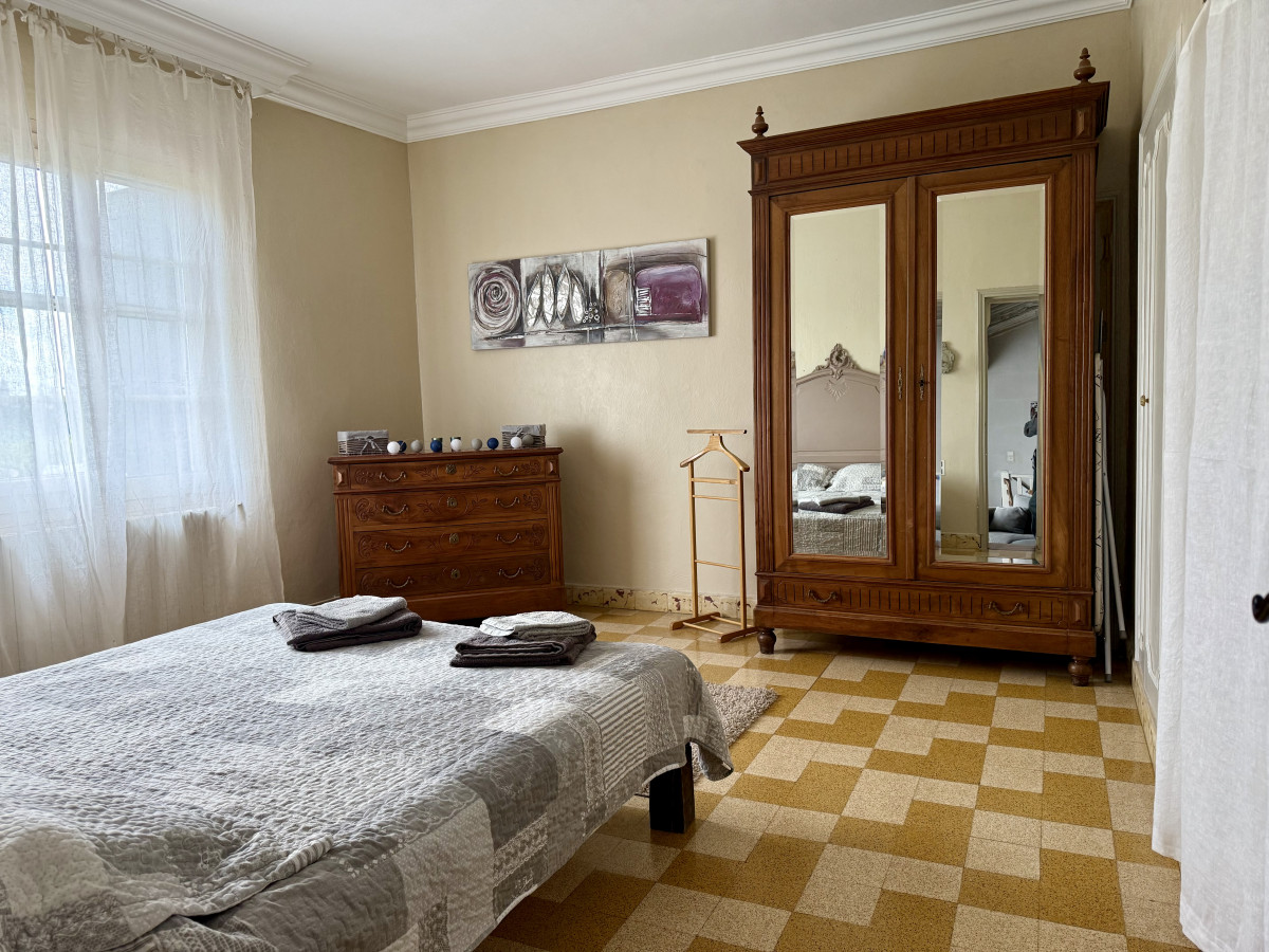 Vue d’ensemble de la chambre du gîte Le Nid d’Amour avec lit double, grande armoire ancienne à portes miroir, commode en bois et sol à carreaux jaunes et blancs.
