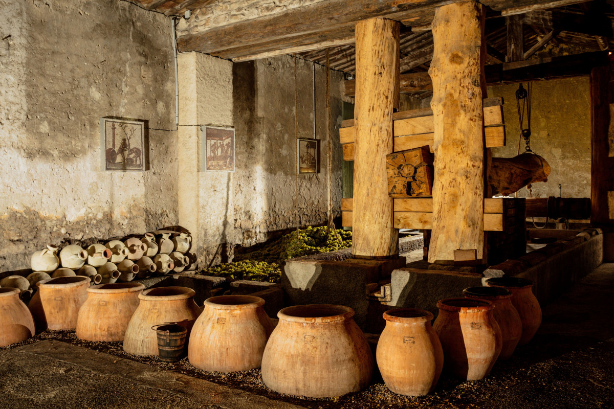 Several terracotta amphorae in the foreground of a building with aged walls, with a wooden wine press installation in the background at Mas des Tourelles in Beaucaire.