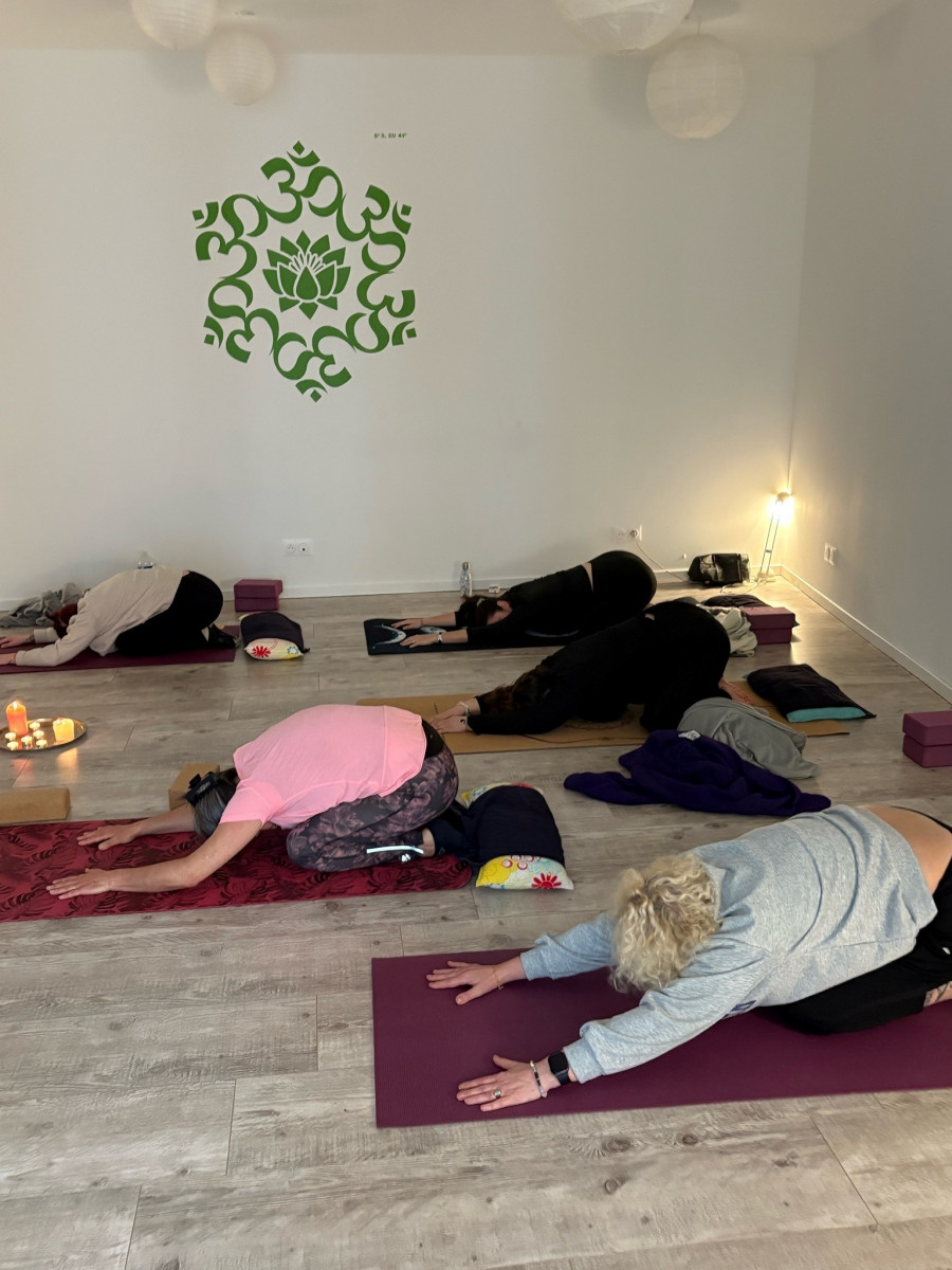 Several participants in a yoga class practising a floor-based relaxation pose, stretching their arms forward on yoga mats in a bright room decorated with yoga symbols.