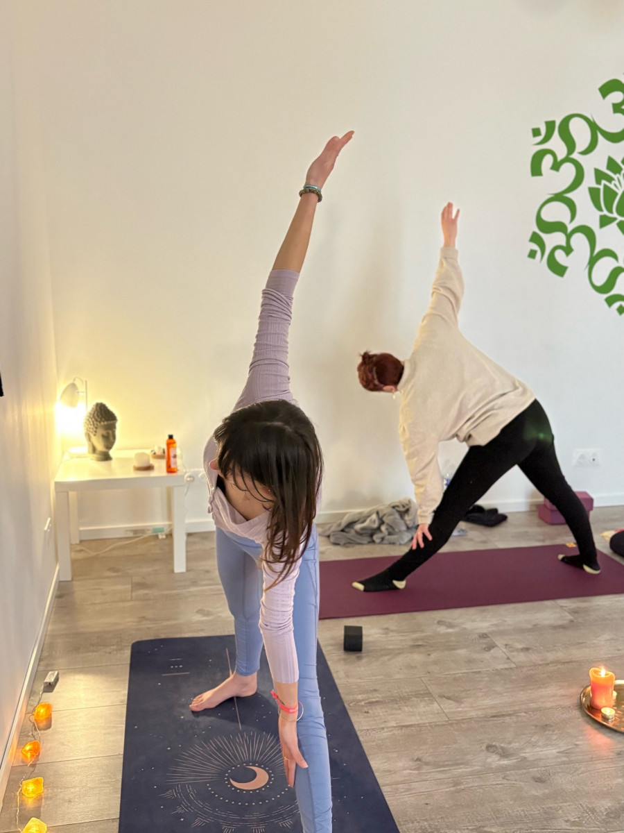 Two participants practising standing yoga poses with one arm raised and the other reaching towards the floor, on yoga mats in a candlelit room.