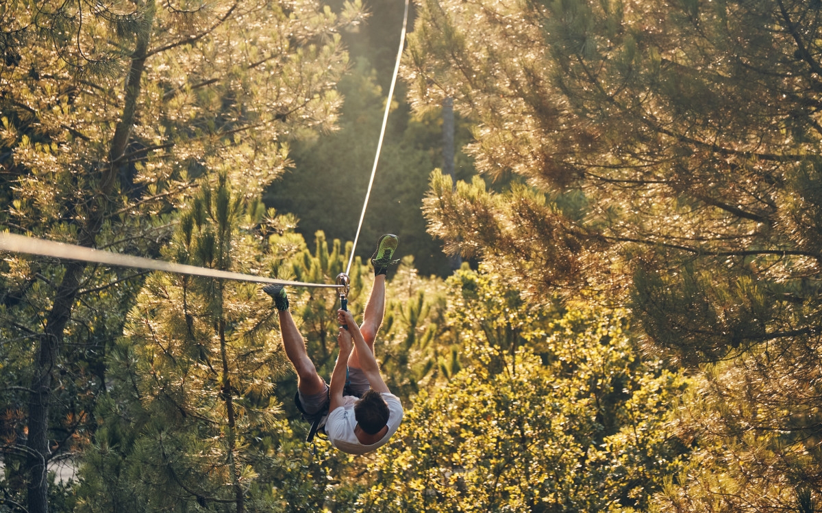 Participant suspended from a rope-based obstacle high above the ground, surrounded by trees and woodland vegetation at the Air de Nature treetop adventure park.