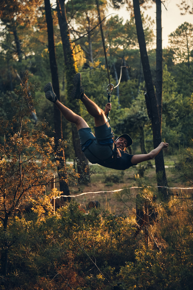 Participant suspended from a zip line, with legs raised and arms outstretched, gliding through a wooded area at the Air de Nature treetop adventure park.