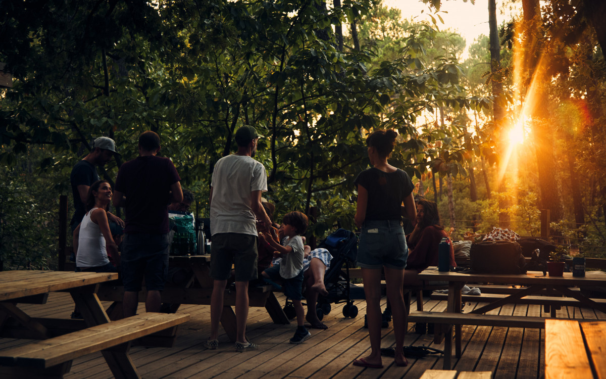 Several visitors gathered on a wooden terrace with tables and benches among the trees at Air de Nature, in the warm late-afternoon sunlight.