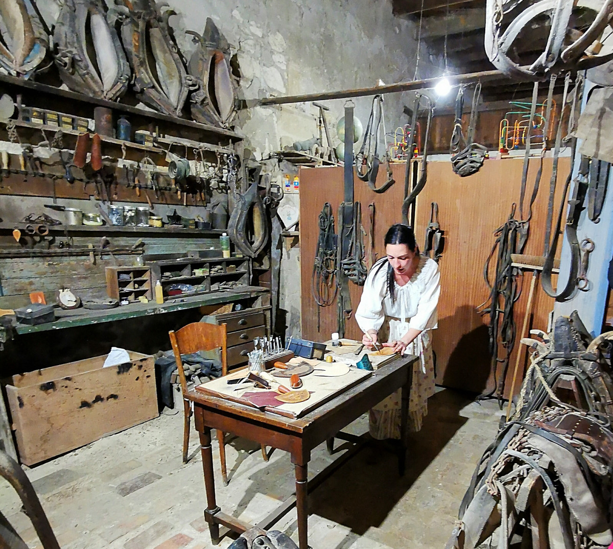 A craftsman works at his bench in a traditional harness-making workshop, surrounded by harnesses, straps and leather equipment hanging on the walls at Le Vieux Mas.