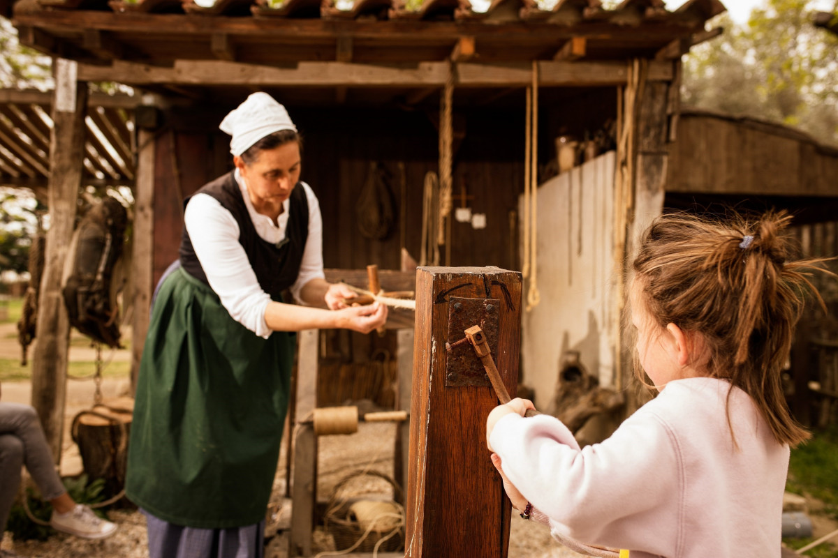 A presenter in traditional costume demonstrates rope making to a child during a workshop on historic crafts at Le Vieux Mas.