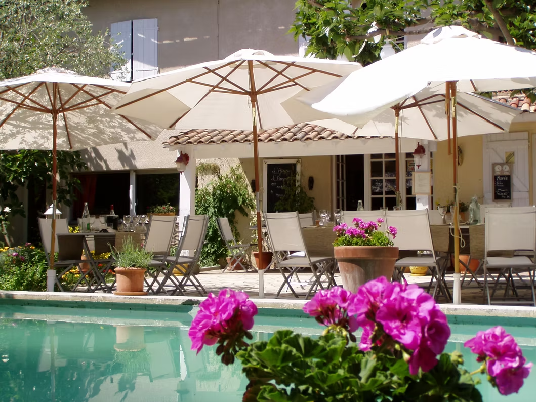 Terrace at Auberge Amandin with tables, chairs and large white parasols beside a swimming pool, with pink flowers in the foreground.