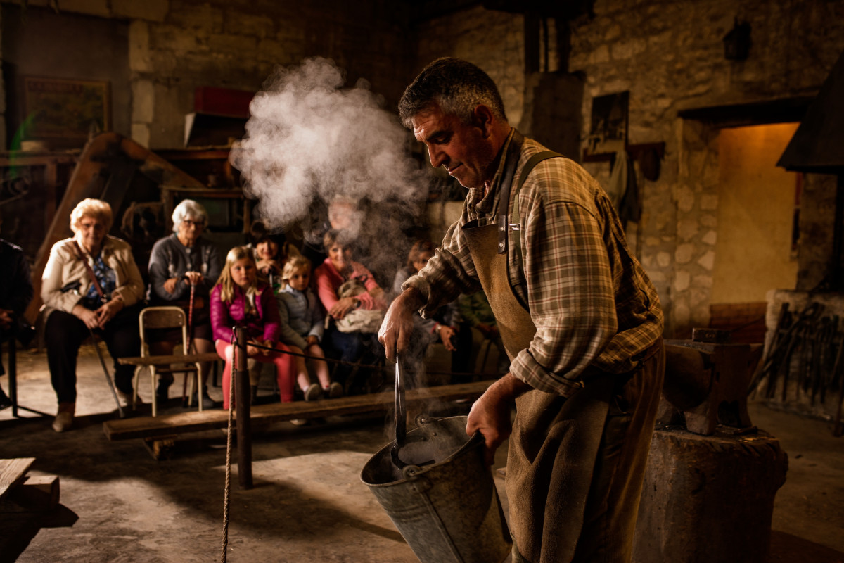 A farrier works in a traditional forge amid smoke, watched by visitors discovering the trade at Le Vieux Mas.