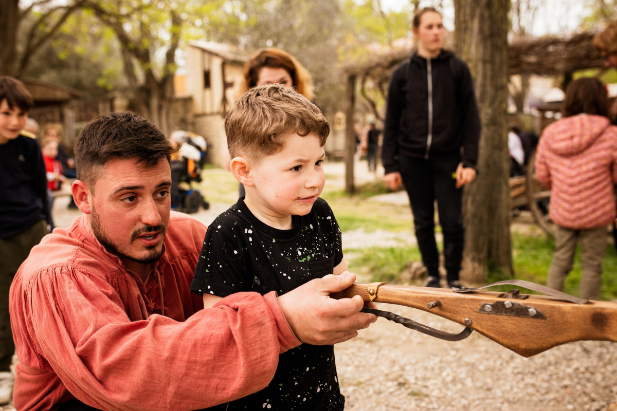 An instructor guides a young boy as he handles a wooden crossbow during a crossbow shooting demonstration at Le Vieux Mas.