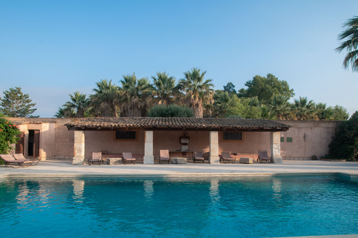 Large outdoor swimming pool at Domaine des Clos, with a stone building providing a shaded seating area and palm trees in the background.