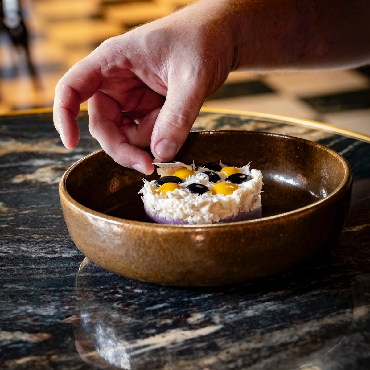 Close-up of a hand adding the finishing touch to a gourmet dish decorated with small yellow and dark elements, served in a deep brown ceramic bowl at Les Vignes Blanches restaurant.