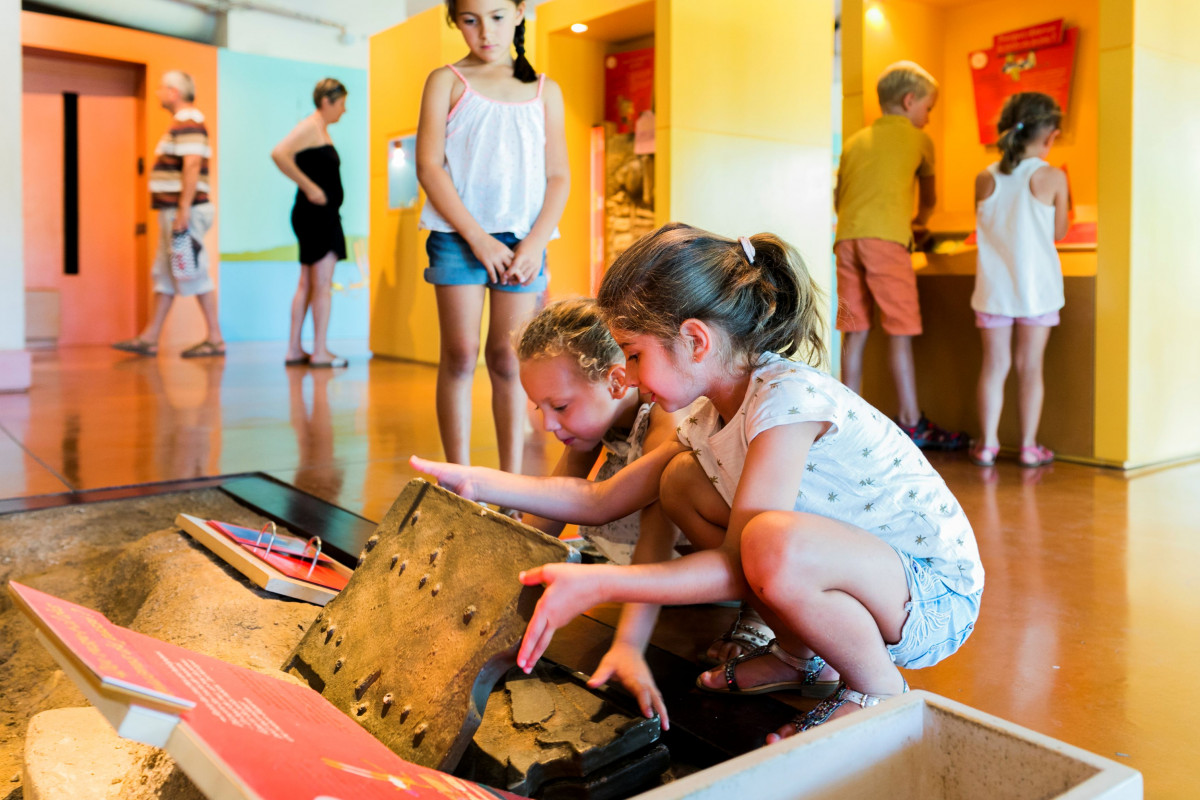 Two children crouching to explore a hands-on educational exhibit in the Pont du Gard Ludo area, while other visitors move around the room.