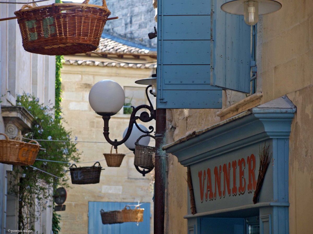 Wicker baskets hanging in a street in Vallabrègues, beside a building with blue shutters and a basket-weaving sign, reflecting the village's traditional craftsmanship.