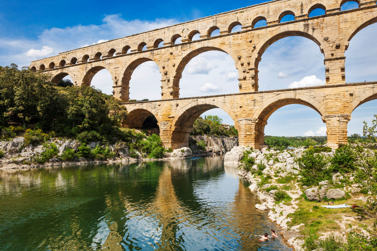 The Pont du Gard and its three tiers of arches seen from the banks of the Gardon River, with water, rocks and vegetation in the foreground.