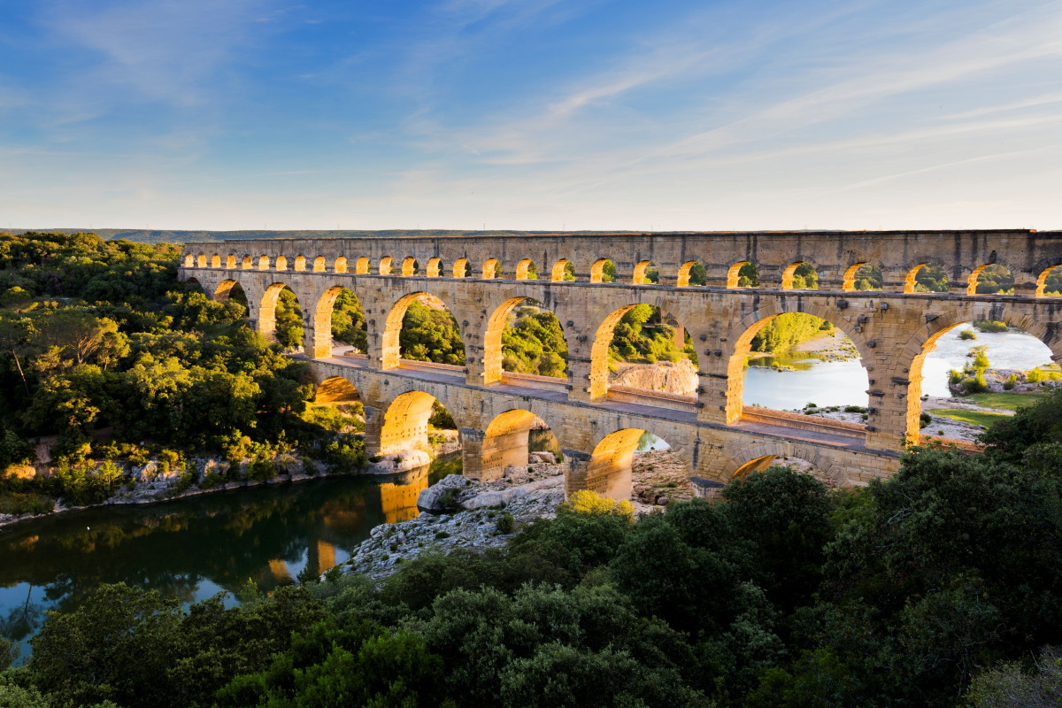 Panoramic view of the Pont du Gard at sunset, with its arches bathed in golden light, the Gardon River and surrounding vegetation.