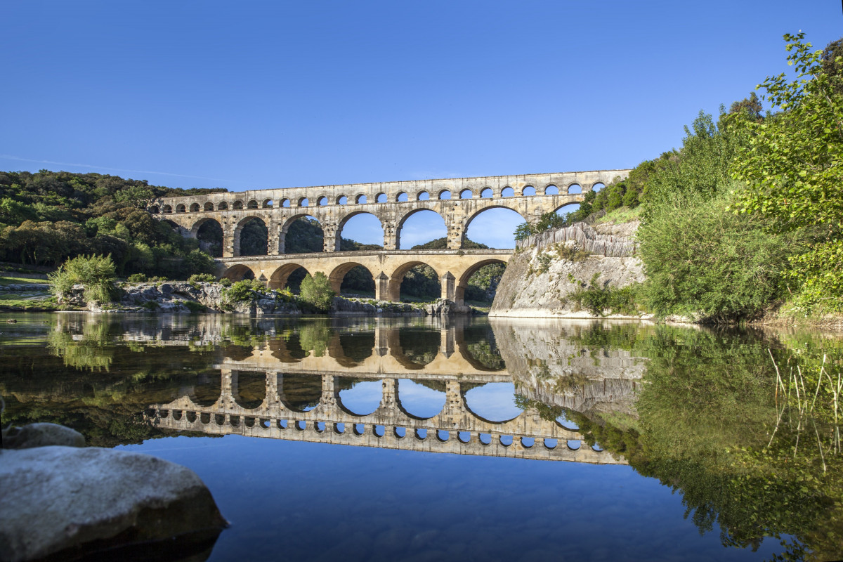 Wide view of the Pont du Gard from the river, with the Roman aqueduct arches reflected in the calm waters of the Gardon and vegetation along the banks.