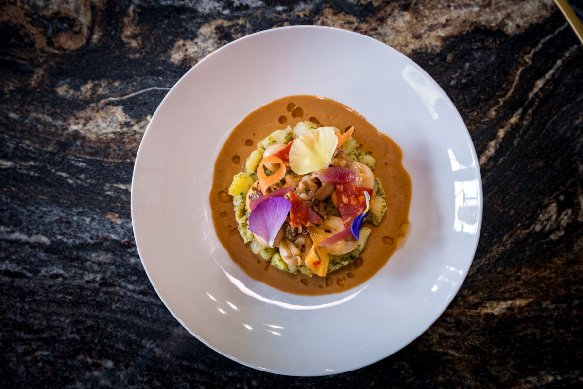 Overhead view of a gourmet dish at Les Vignes Blanches restaurant, featuring a colourful central preparation with vegetables, edible flowers and brown sauce, served on a large white plate.