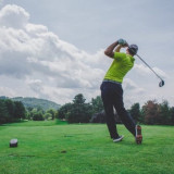 Golfer wearing a green shirt completing a swing on the Golf de Saint-Roman course, with green grass, trees and hills in the background.