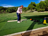 Young golfer practising a golf shot on a driving-range mat at Golf de Saint-Roman, with training equipment and a tree-lined course in the background.