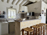 Vue de la cuisine ouverte du gîte Le Nid d’Amour, avec comptoir habillé de pierre, tabourets hauts en bois, électroménager et poutres apparentes.