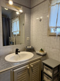 Vue de la salle de bains du gîte Le Nid d’Amour avec vasque blanche, grand miroir éclairé, meuble de rangement et fenêtre à rideau blanc.