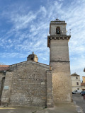 Exterior view of the church in Fourques, showing its stone façade, tall square bell tower and bell structure against a cloudy sky.