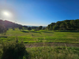 Wide panoramic view of Golf de Saint-Roman in low sunlight, featuring green fairways, trees and wooded hills.