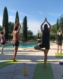 Participants practising yoga on mats beside the swimming pool at Domaine des Clos, balancing on one leg with their hands joined above their heads.