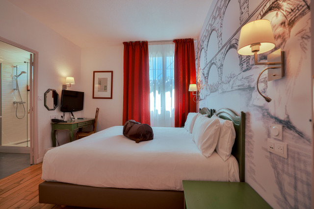 Hôtel Les Vignes Blanches – Double Bedroom Guest room at Hôtel Les Vignes Blanches with a white double bed, red curtains, wall lights and a decorative wall design.