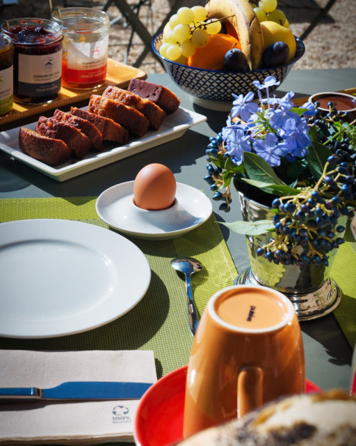 Domaine des Clos – Breakfast Table Breakfast table at Domaine des Clos with slices of cake, fresh fruit, a boiled egg, jams, a cup and a bouquet of blue flowers.