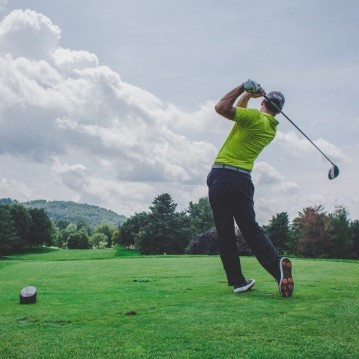 Golfer wearing a green shirt completing a swing on the Golf de Saint-Roman course, with green grass, trees and hills in the background.