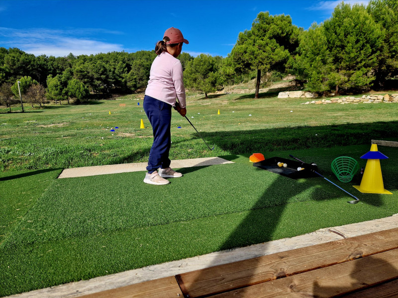 Golfer standing on a driving-range mat at Golf de Saint-Roman, holding a club while facing the green course, with training equipment nearby.