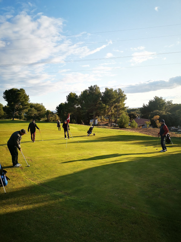 Several golfers spread across a sunlit green at Golf de Saint-Roman, practising putting in a green, tree-lined setting.
