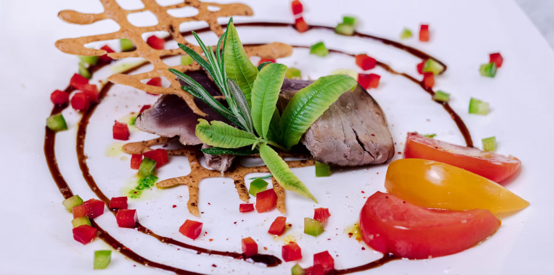 Assiette gastronomique aux tomates – Auberge Amandin Assiette gastronomique de l'Auberge Amandin présentant une pièce de viande tranchée, des tomates rouges et jaunes, des herbes fraîches et une sauce disposée en cercle.