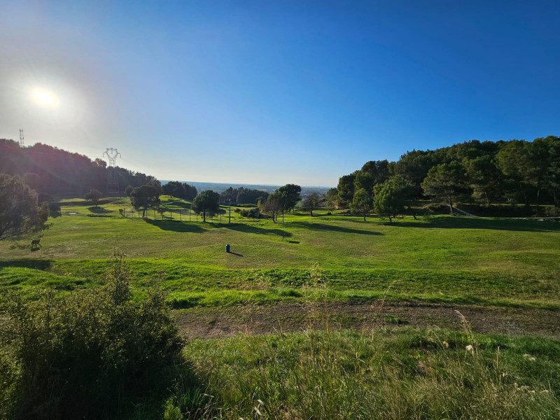 Wide panoramic view of Golf de Saint-Roman in low sunlight, featuring green fairways, trees and wooded hills.