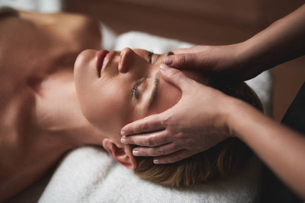 Close-up of a person lying down with their eyes closed, receiving a head massage, with the practitioner's hands resting on the temples and sides of the head.